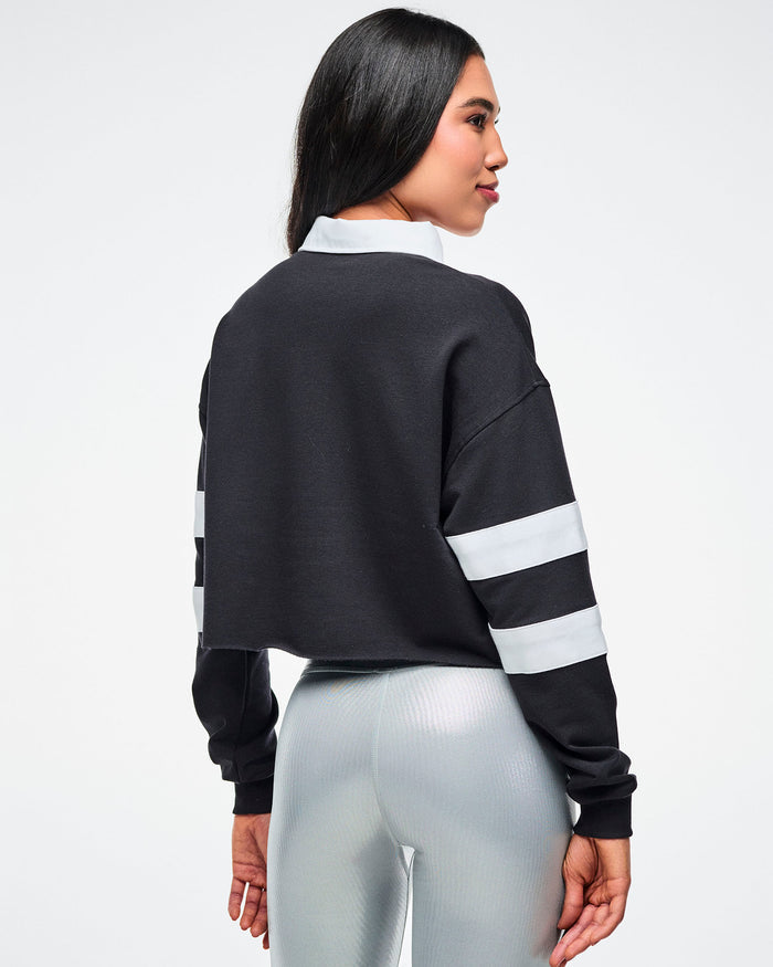 Back view of a female model wearing a black cropped polo with white sleeve stripes, a white collar and paired with silver biker shorts.