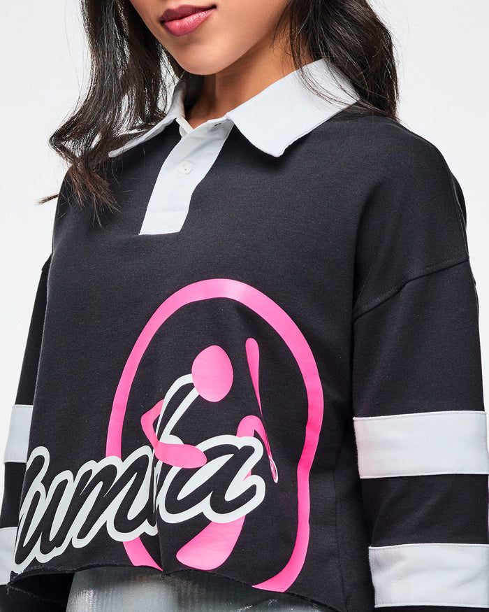 Close view of the black cropped polo with bold white “ZUMBA” text and a large pink Zumbito graphic, showcasing the white collar and white sleeve stripes.