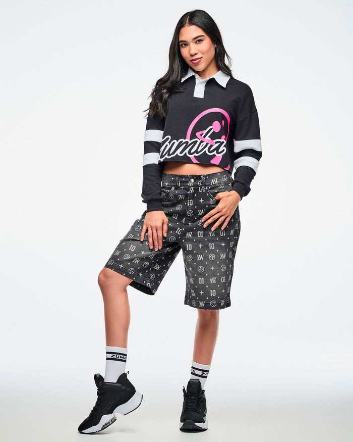 Female model in a black-and-white striped cropped polo featuring bold white “ZUMBA” text and a large pink Zumbito graphic, paired with patterned shorts plus black-and-white socks and sneakers.