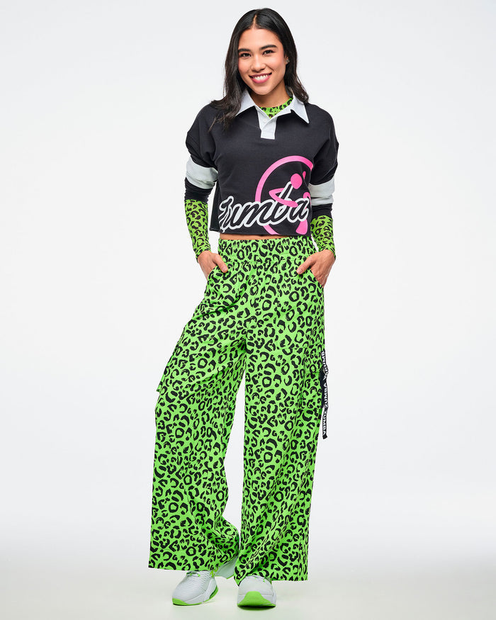 Female model in a black‑and‑white striped cropped polo with bold white “ZUMBA” text and a large pink Zumbito graphic, paired with neon green leopard‑print cargo pants and white‑and‑green sneakers.