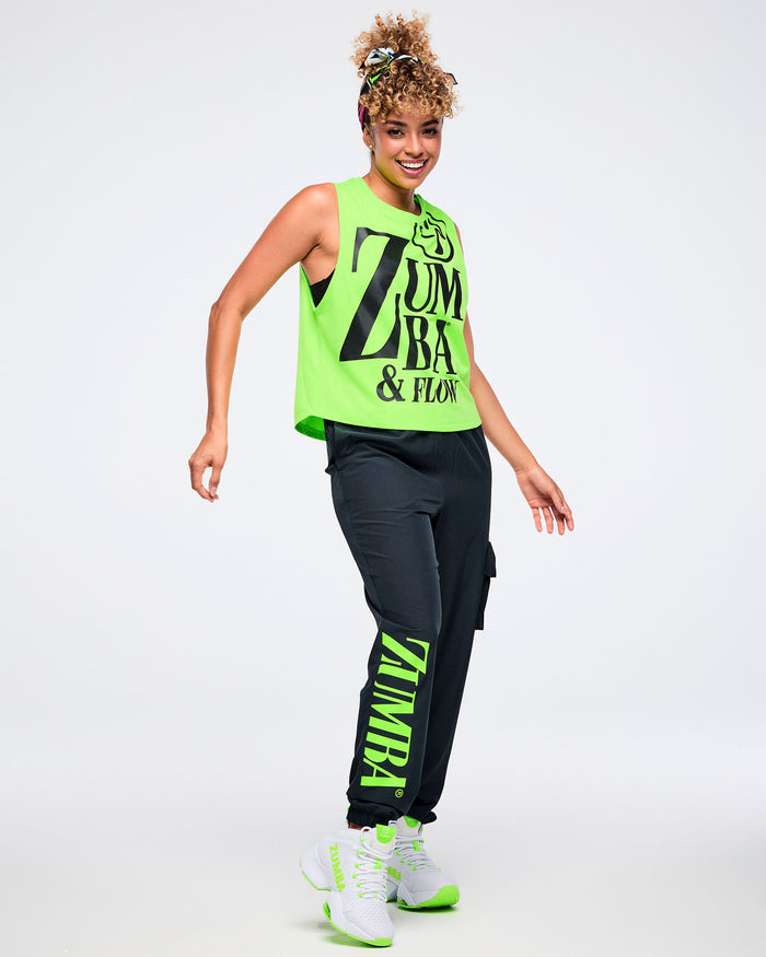 Full view of a female model wearing a neon green sleeveless Zumba tank with oversized black “ZUMBA & FLOW” text and Zumbito graphic, paired with black pants and green and white shoes.