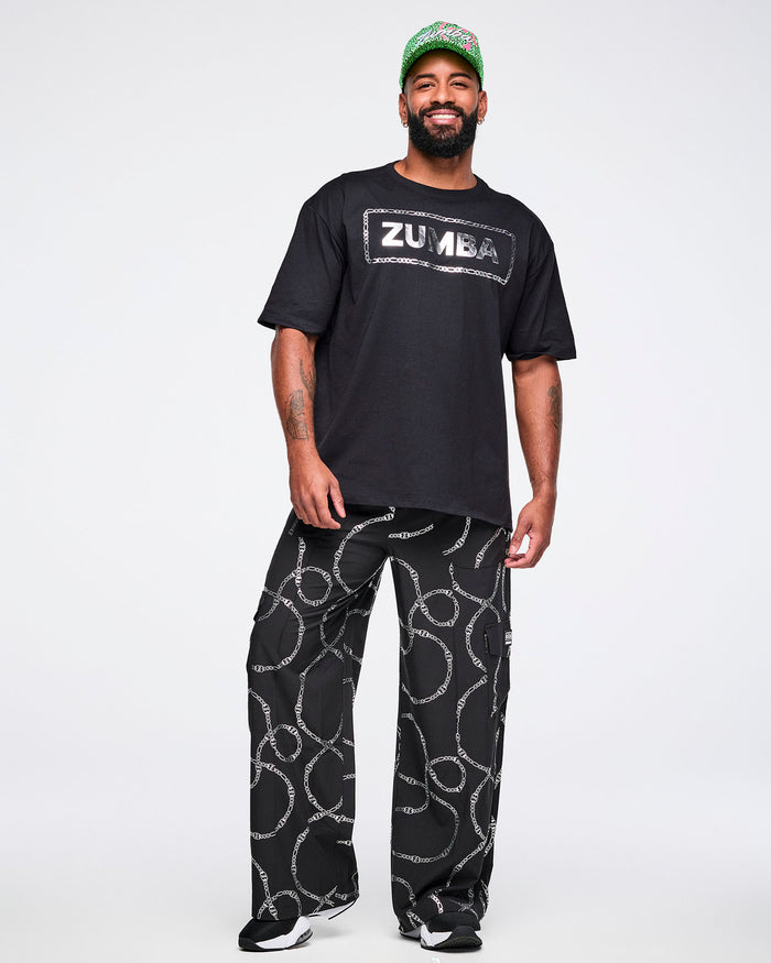 Full view of a male model wearing a black Zumba T-shirt with “ZUMBA” in a silver patterned rectangle, paired with matching pants and black and white shoes.