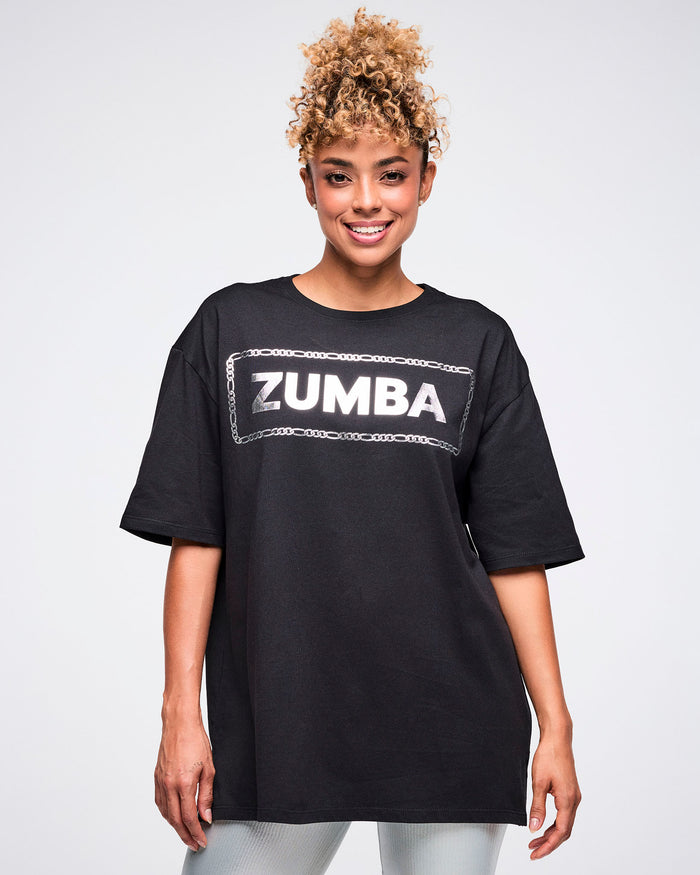 Female model wearing a black Zumba T-shirt with “ZUMBA” in a silver patterned rectangle, paired with matching pants.