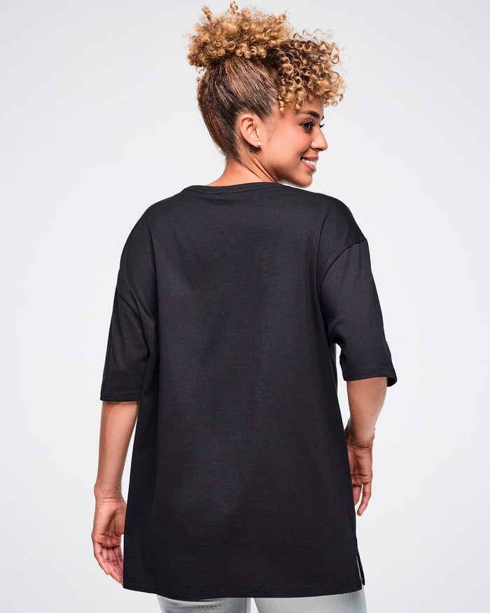 Back view of a female model wearing a black Zumba T-shirt with “ZUMBA” in a silver patterned rectangle, paired with matching pants.