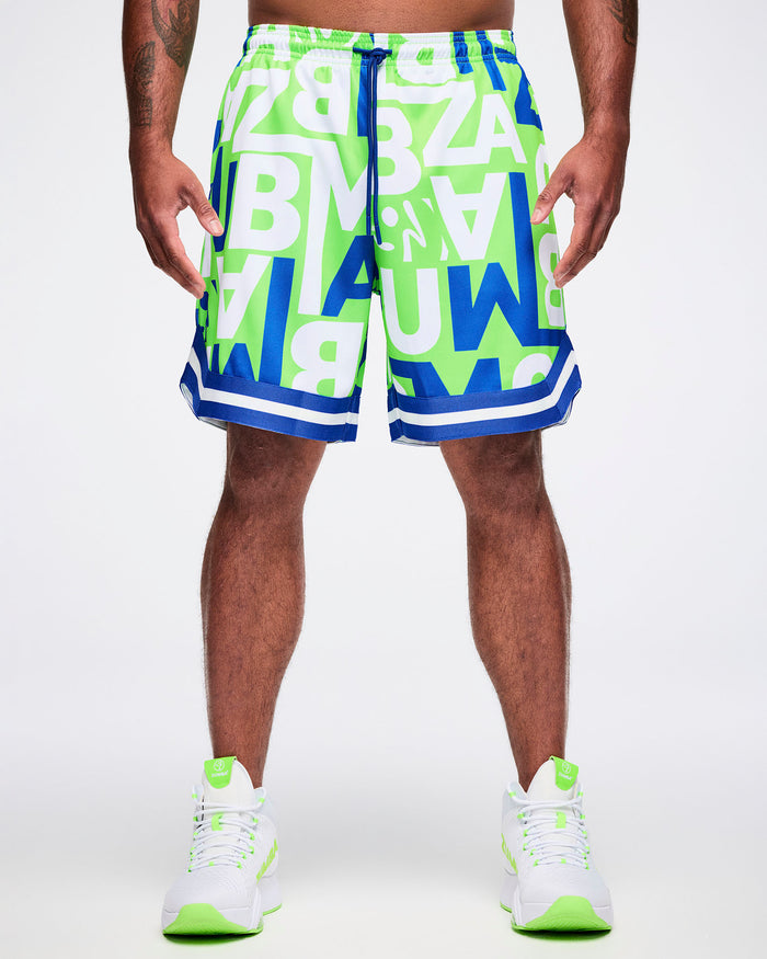 Front view of a male model wearing green, blue and white patterned ZUMBA lettering shorts paired with green and white shoes.