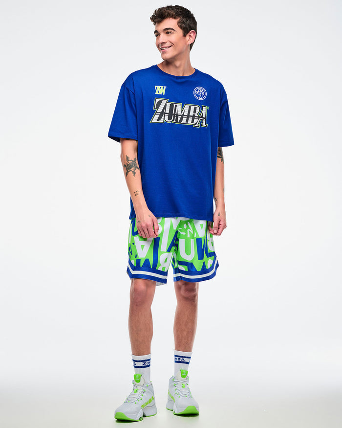 Full view of a male model wearing green, blue, and white patterned shorts with ZUMBA lettering, paired with a blue top, blue-and-white socks, and green-and-white shoes.