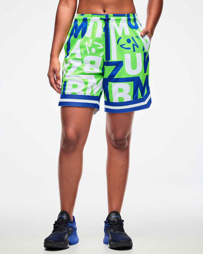 Female model wearing green, blue, and white patterned shorts with ZUMBA lettering, one hand in her pocket, paired with blue-and-black shoes.