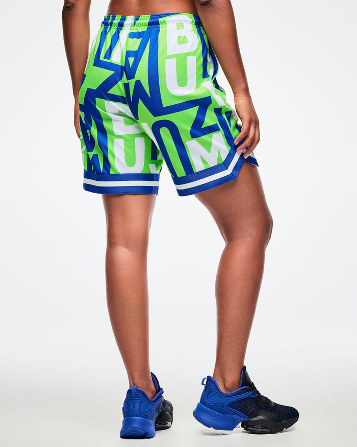 Back view of a female model wearing green, blue, and white patterned shorts with ZUMBA lettering, one hand in her pocket, paired with blue-and-black shoes.