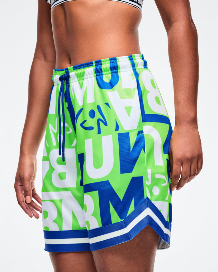 Close-up view of a female model wearing green, blue, and white patterned shorts with ZUMBA lettering, one hand in her pocket, paired with blue-and-black shoes.