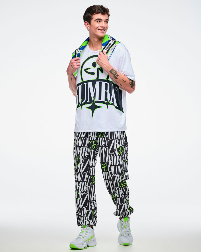 Male model holds a colorful bandana at his neck and wears a white Zumba top with a black and green logo, black‑white‑green pants, and white‑green shoes on a white background.
