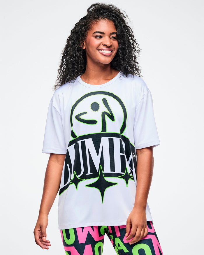 Female model in a white Zumba top with a black and green logo and paired with multicolored leggings.