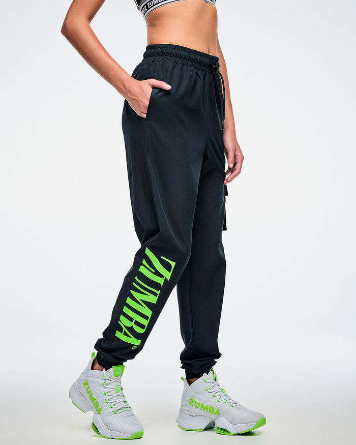 Side view of a female model with her right hand in the pocket of black cargo pants featuring green “ZUMBA” text on the right leg, paired with green-and-white shoes.