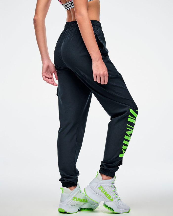 Back view of a female model wearing black cargo pants featuring green “ZUMBA” text on the right leg, paired with green-and-white shoes.