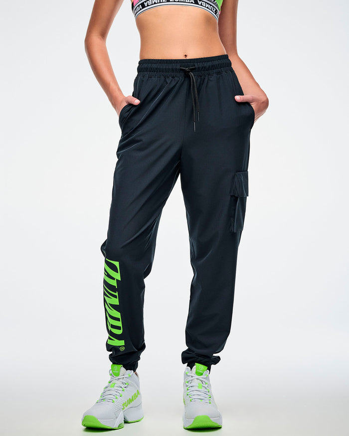 Front view of a female model with both hands in the pocket of black cargo pants featuring green “ZUMBA” text on the right leg, paired with green-and-white shoes.