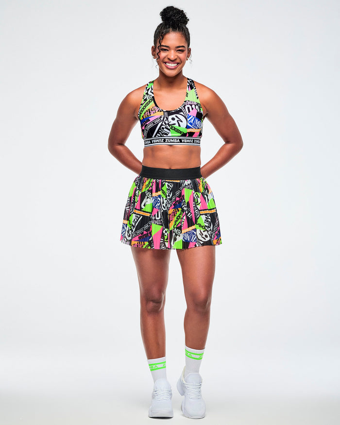 Full view of a female model wearing a skort with a vibrant graffiti-style print in neon green, pink, yellow, and black, paired with green and white socks and white sneakers.