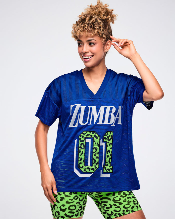 Female model wearing a blue Zumba mesh jersey with white “ZUMBA” text and a green leopard-print “01,” paired with matching green biker shorts.