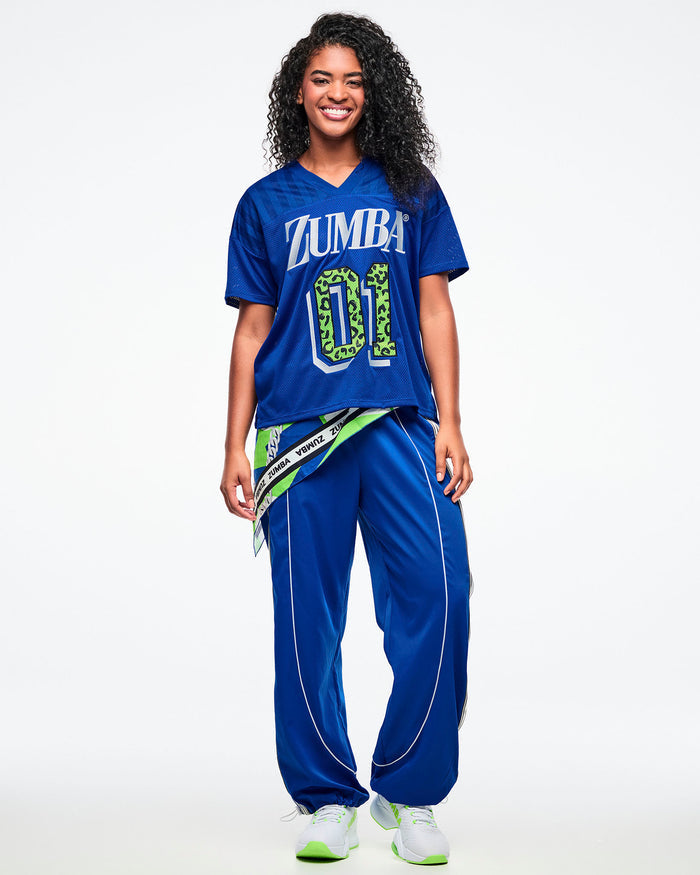 Female model wearing a blue Zumba mesh jersey with white “ZUMBA” text and a green leopard-print “01,” paired with blue-and-white pants, green-and-white shoes, and a bandana tied around her waist.