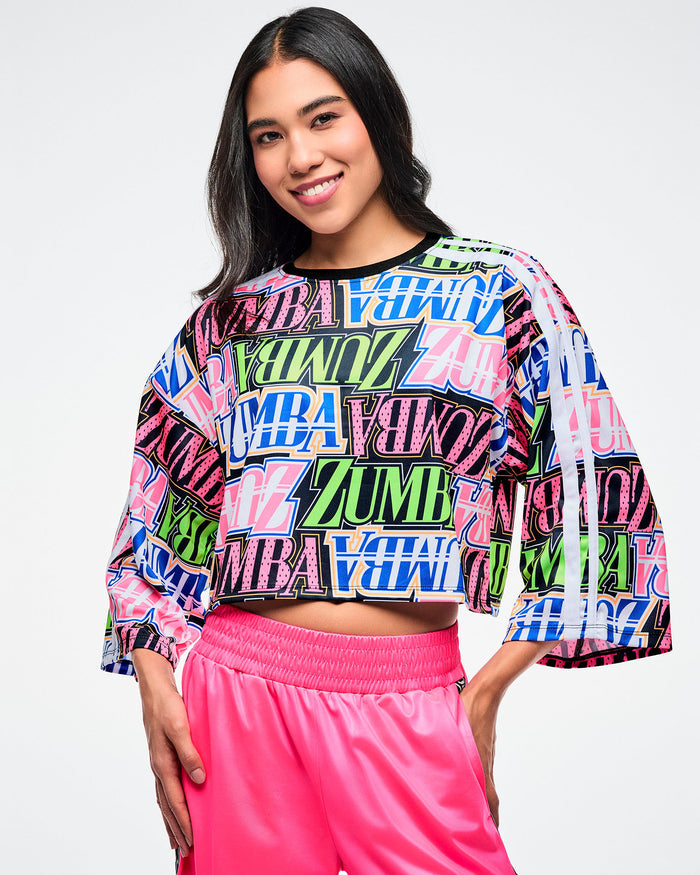 Female model wearing a colorful Zumba boxy crop top and pink pants on a white background.