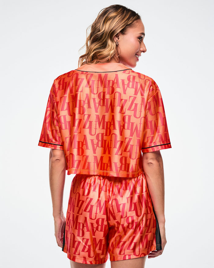 Back view of a female model wearing an orange baseball top with red "ZUMBA" patterned text paired with matching boxer shorts on a white background.