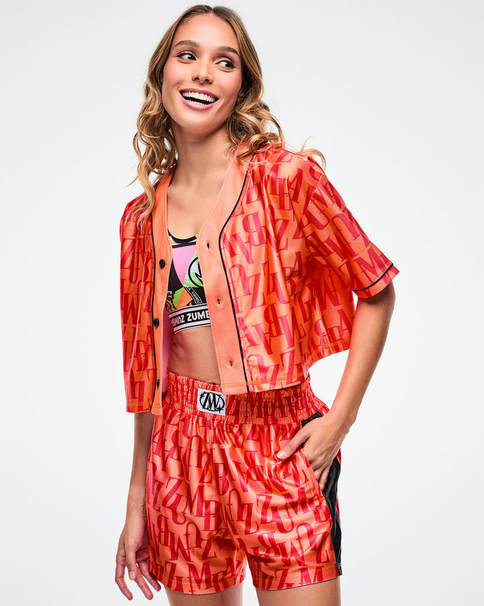 Female model in an unbuttoned orange baseball top with red “ZUMBA” text, layered over a multicolored Zumba bra, one hand in her pocket.