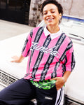 Female model wearing a pink-and-black striped Zumba top with white cursive “Zumba,” layered over a green mesh collared polo, paired with black-and-white striped track pants, leaning against the hood of a white car.