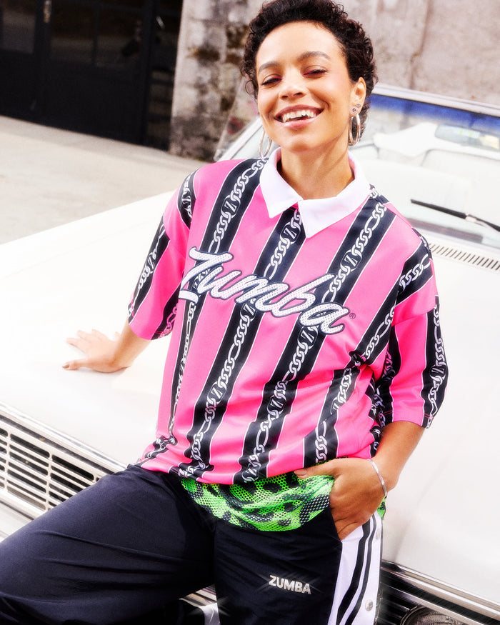 Female model wearing a pink-and-black striped Zumba top with white cursive “Zumba,” layered over a green mesh collared polo, paired with black-and-white striped track pants, leaning against the hood of a white car.