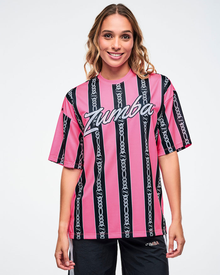 Female model wearing a pink-and-black striped Zumba top with white cursive “Zumba,” and black and white striped track pants on a white background.