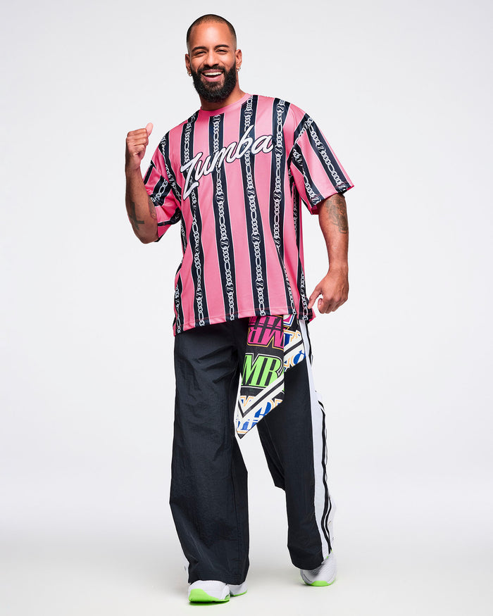 Male model wearing a pink-and-black striped Zumba top with white cursive “Zumba,” black-and-white striped pants, green-and-white shoes, and a bandana tucked into his pants.