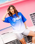 Female model wearing a blue-to-white gradient Zumba tee paired with silver biker shorts, posing on the hood of a white car.