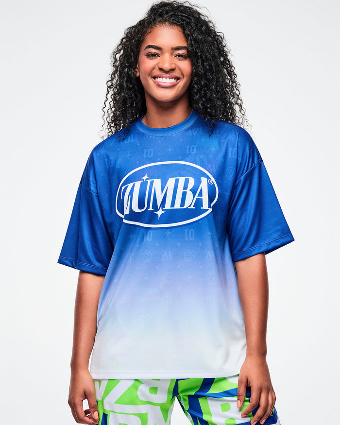 Female model wearing a blue-to-white gradient Zumba tee paired with blue and green patterned Zumba pants.