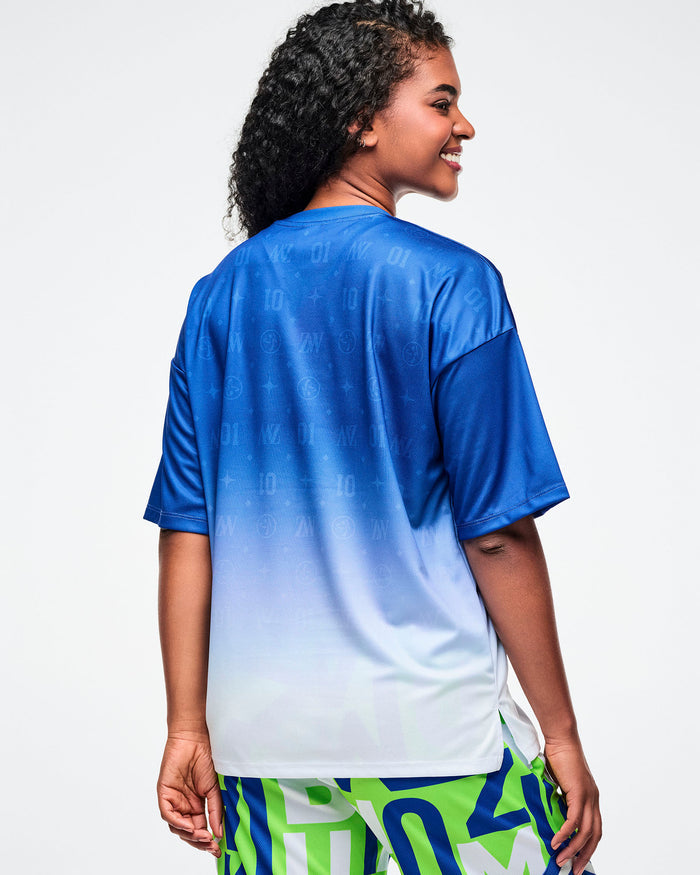 Back view of a female model wearing a blue-to-white gradient Zumba tee paired with blue and green patterned Zumba pants.