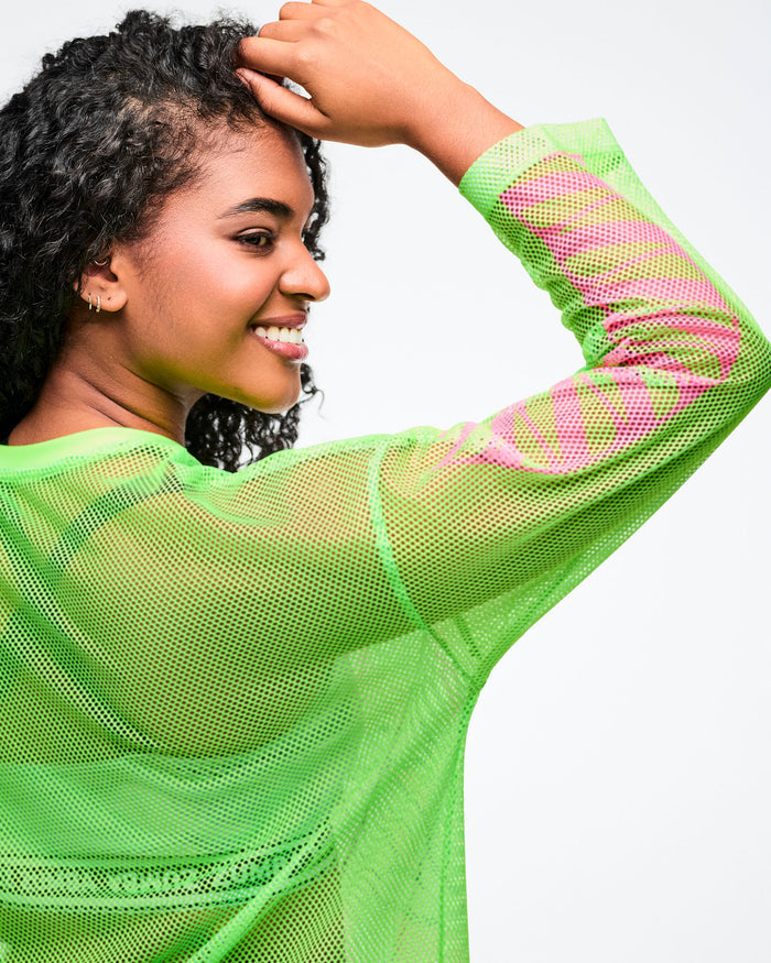 Close-up of a female model wearing a neon green mesh long‑sleeve Zumba top with pink sleeve accents, layered over a silver bra.