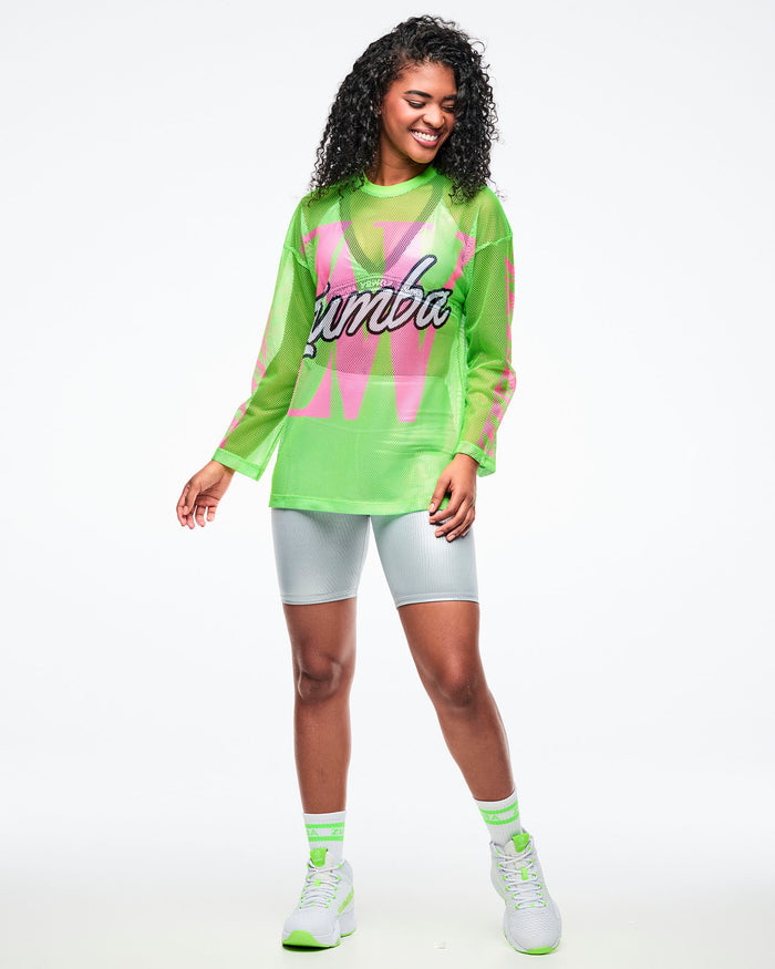 Female model wearing a neon green mesh long-sleeve Zumba top with pink sleeve accents, layered over a silver bra and paired with matching silver biker shorts, plus green-and-white socks and shoes.