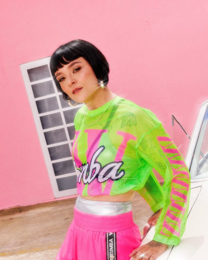 Female model wearing a neon green mesh top with pink and white graphic lettering, paired with pink sweatpants, leaning against a white car.
