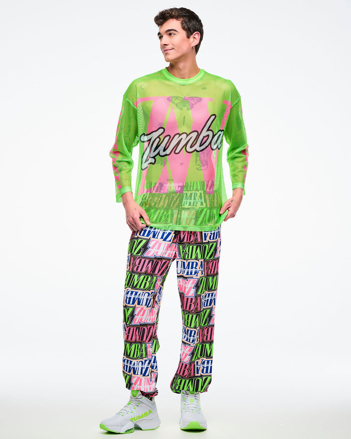 Male model wearing a neon green mesh long-sleeve Zumba top with bold pink graphics and white cursive “Zumba,” paired with multicolored Zumba pants and green-and-white shoes.