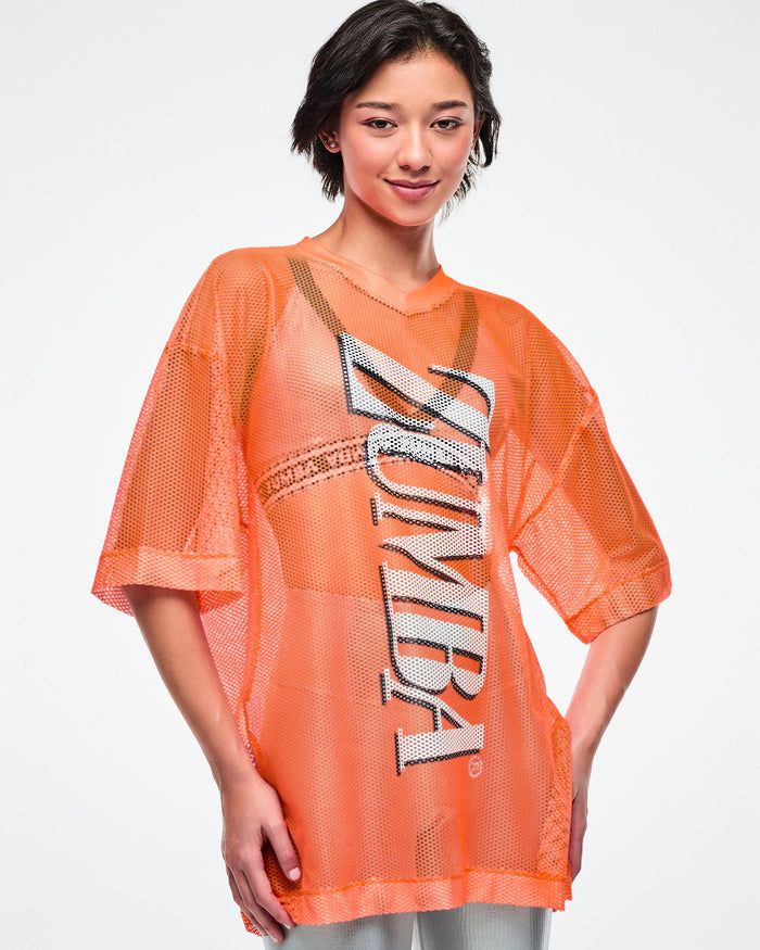 Female model wearing an oversized orange mesh Zumba top with bold vertical “ZUMBA” silver lettering paired with silver biker shorts and a silver bra underneath.