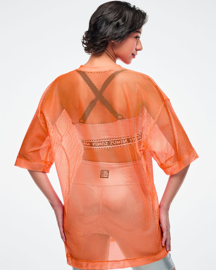 Back view of a female model wearing an oversized orange mesh Zumba top, paired with silver biker shorts and silver bra.