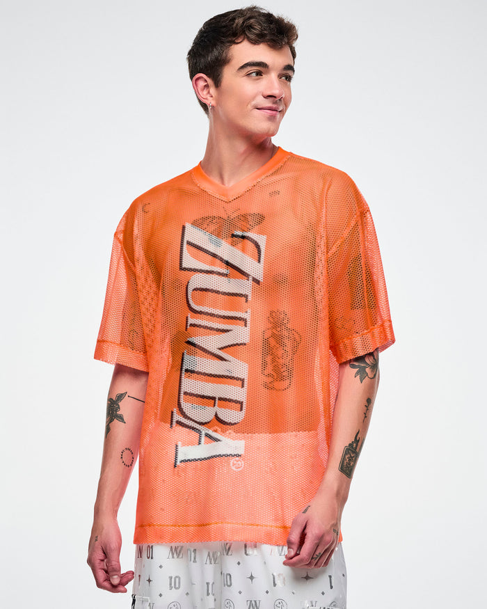 Male model wearing an oversized orange mesh Zumba top with bold vertical “ZUMBA” silver lettering paired with white and silver pants.