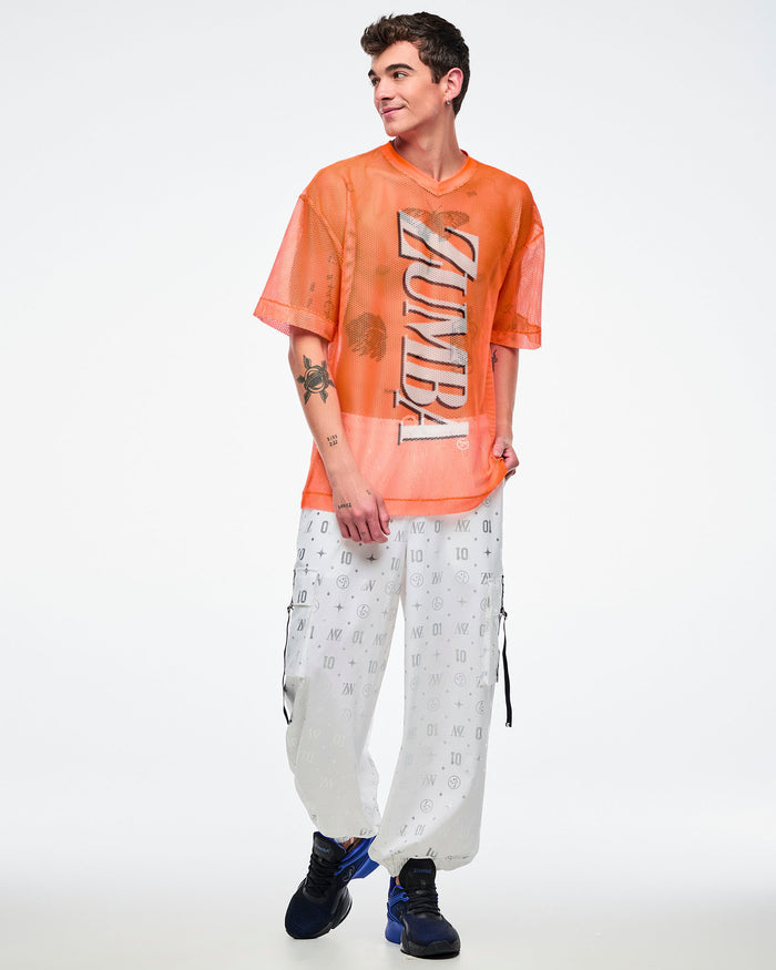 Male model wearing an oversized orange mesh Zumba top with bold vertical “ZUMBA” silver lettering paired with white and silver pants and blue and black shoes.
