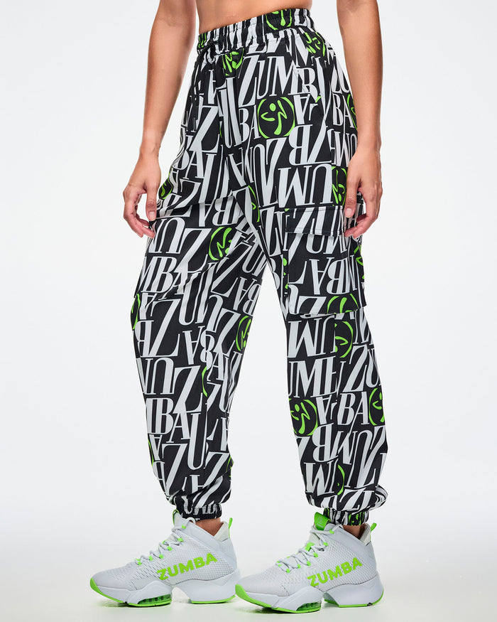 Female model wearing black and white patterned ZUMBA pants with green Zumbito accents, paired with white and green sneakers on a white background.