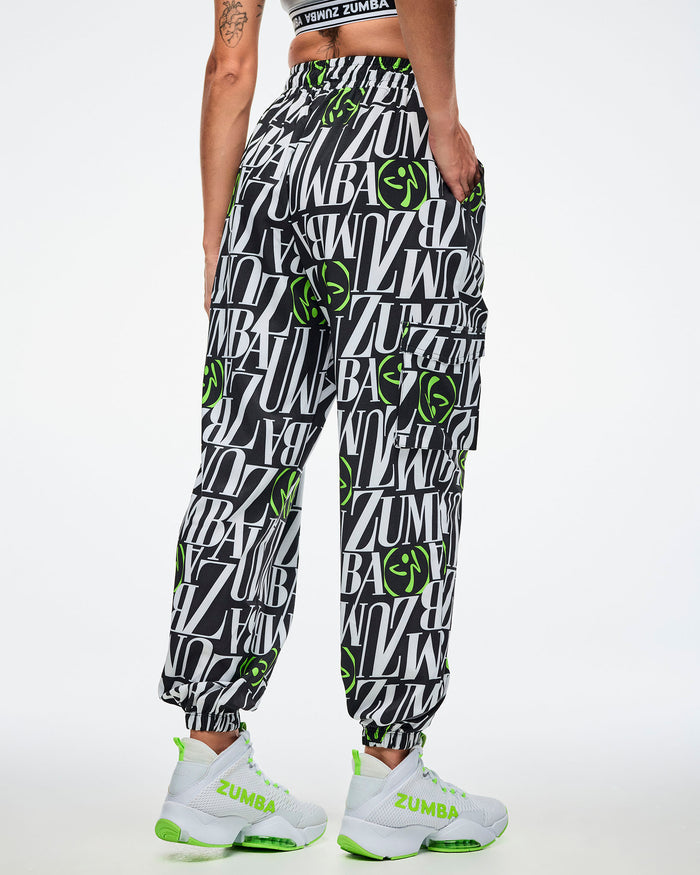 Back view of a female model wearing black and white patterned ZUMBA pants with green Zumbito accents, paired with white and green sneakers on a white background.