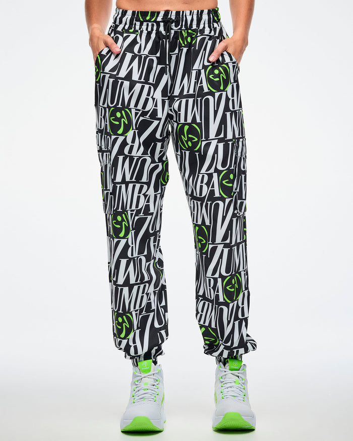Front view of a female model with hands in her pockets, wearing black and white patterned ZUMBA pants with green Zumbito accents, paired with white and green sneakers on a white background.