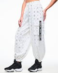 Female model in white cargo pants printed with silver Zumba motifs (ZW, Zumbito, dots, stars, “01”), featuring a black‑and‑white ribbon on the cargo pocket reading “Zumba,” in white, paired with black‑and‑white shoes.