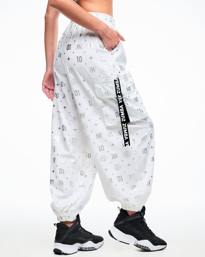 Back view of a female model in white cargo pants printed with silver Zumba motifs (ZW, Zumbito, dots, stars, “01”), featuring a black‑and‑white ribbon on the cargo pocket reading “Zumba,” in white, paired with black‑and‑white shoes.