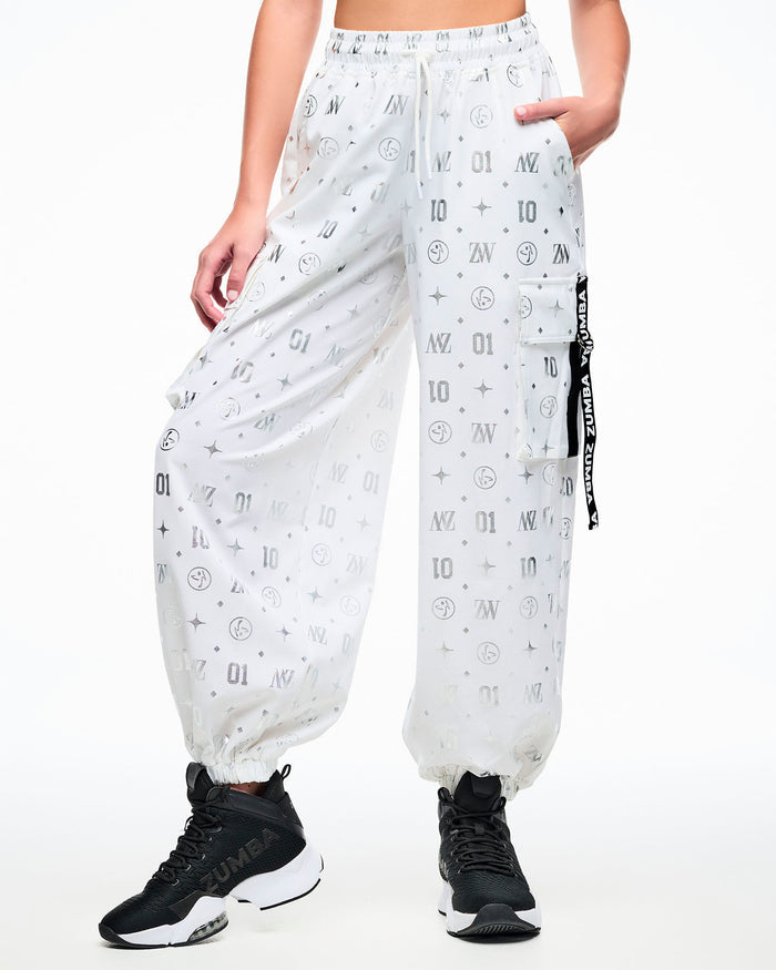 Front view of a female model in white cargo pants printed with silver Zumba motifs (ZW, Zumbito, dots, stars, “01”), featuring a black‑and‑white ribbon on the cargo pocket reading “Zumba,” in white, paired with black‑and‑white shoes.
