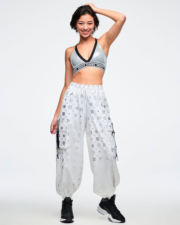 Female model in a silver sports bra and white cargo pants featuring a silver Zumba pattern paired with black and white shoes.