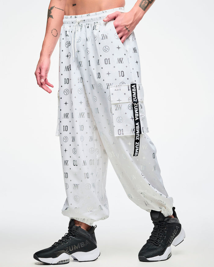 Male model in white cargo pants printed with silver Zumba motifs (ZW, Zumbito, dots, stars, “01”), featuring a black‑and‑white ribbon on the cargo pocket reading “Zumba,” in white, paired with black‑and‑white shoes.