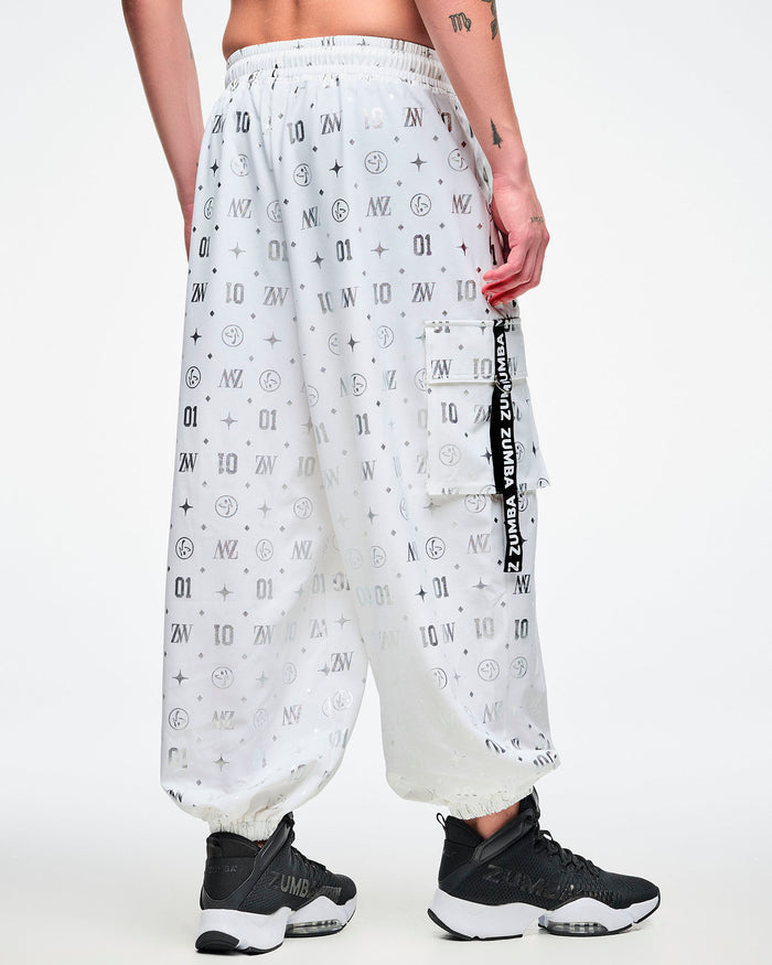 Back view of a male model in white cargo pants printed with silver Zumba motifs (ZW, Zumbito, dots, stars, “01”), featuring a black‑and‑white ribbon on the cargo pocket reading “Zumba,” in white, paired with black‑and‑white shoes.