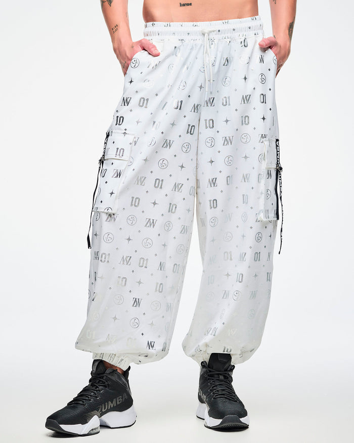 Front view of a male model in white cargo pants printed with silver Zumba motifs (ZW, Zumbito, dots, stars, “01”), featuring a black‑and‑white ribbon on the cargo pocket reading “Zumba,” in white, paired with black‑and‑white shoes.