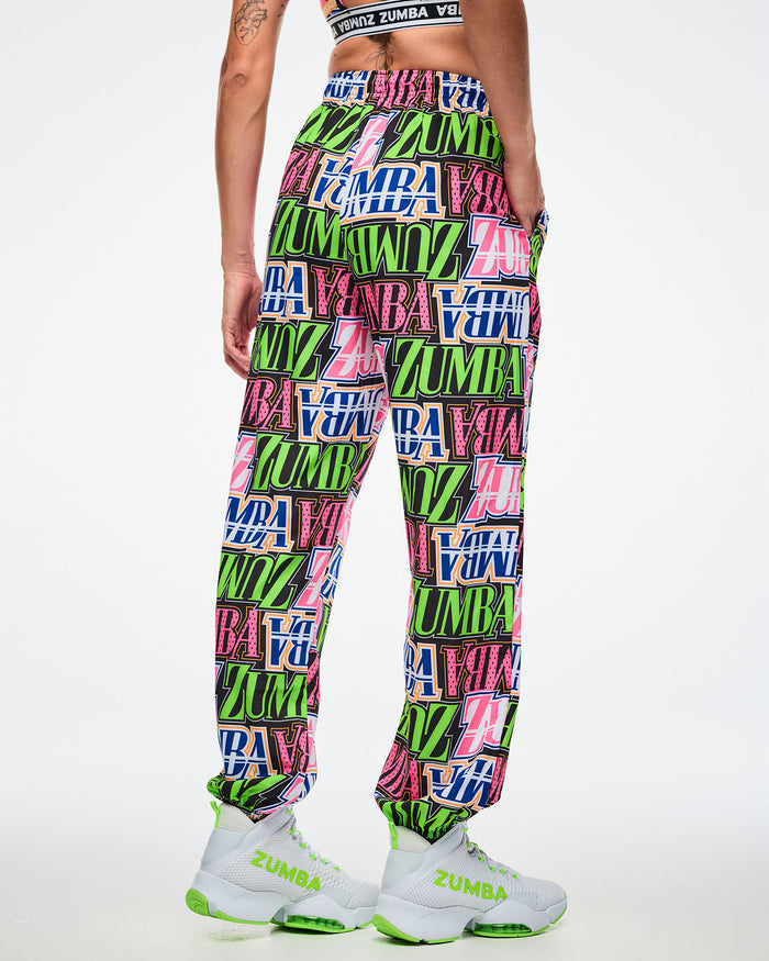 Back view of a female model wearing colorful Zumba pants paired with green and white shoes on a white background.
